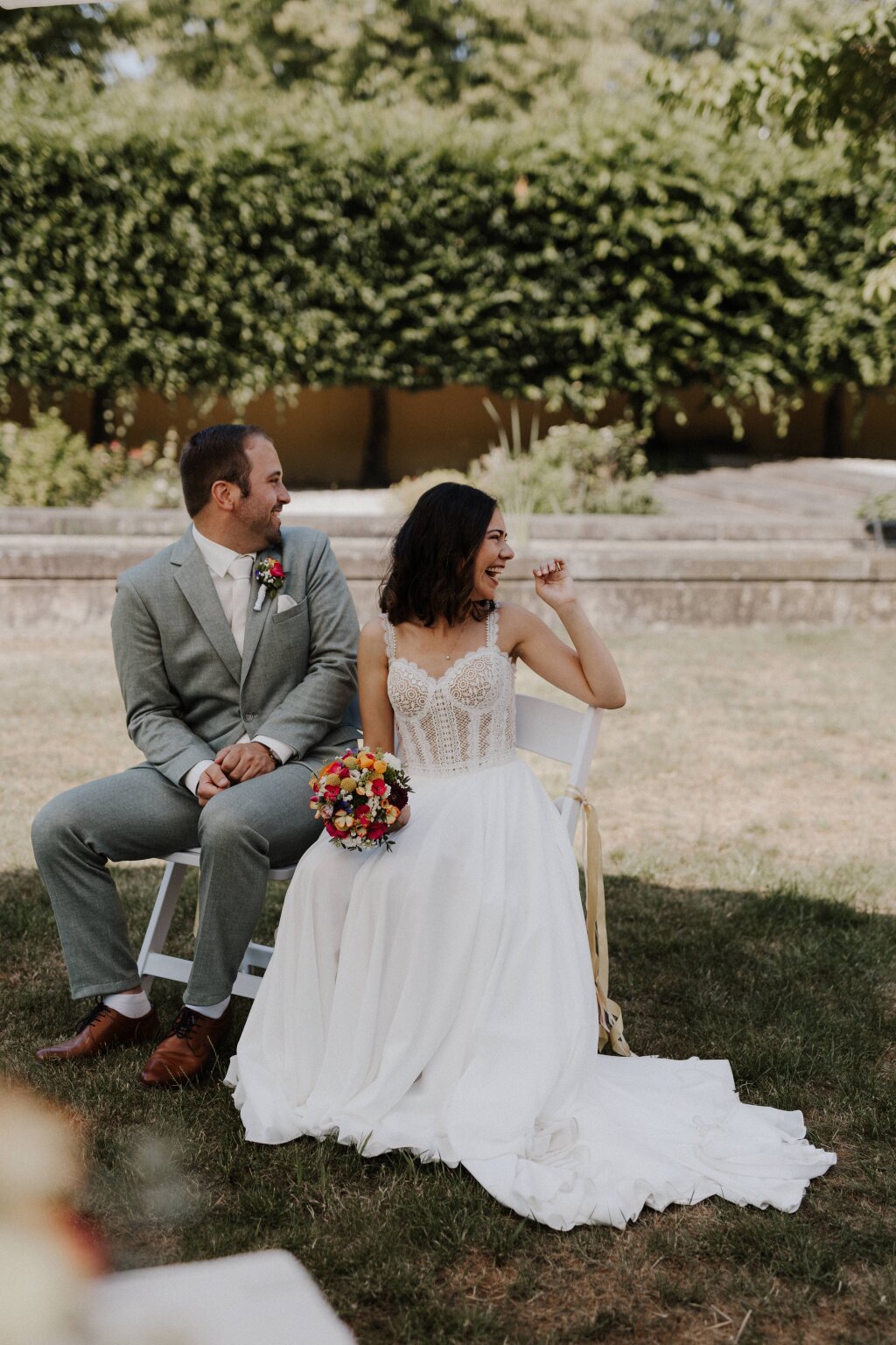 Lachendes Brautpaar während der Trauung auf der Wiese. 
Die Braut hat einen schönen bunten Strauß in der Hand. Im Vordergrund ist ein Teil des Trautisches zu erkennen.