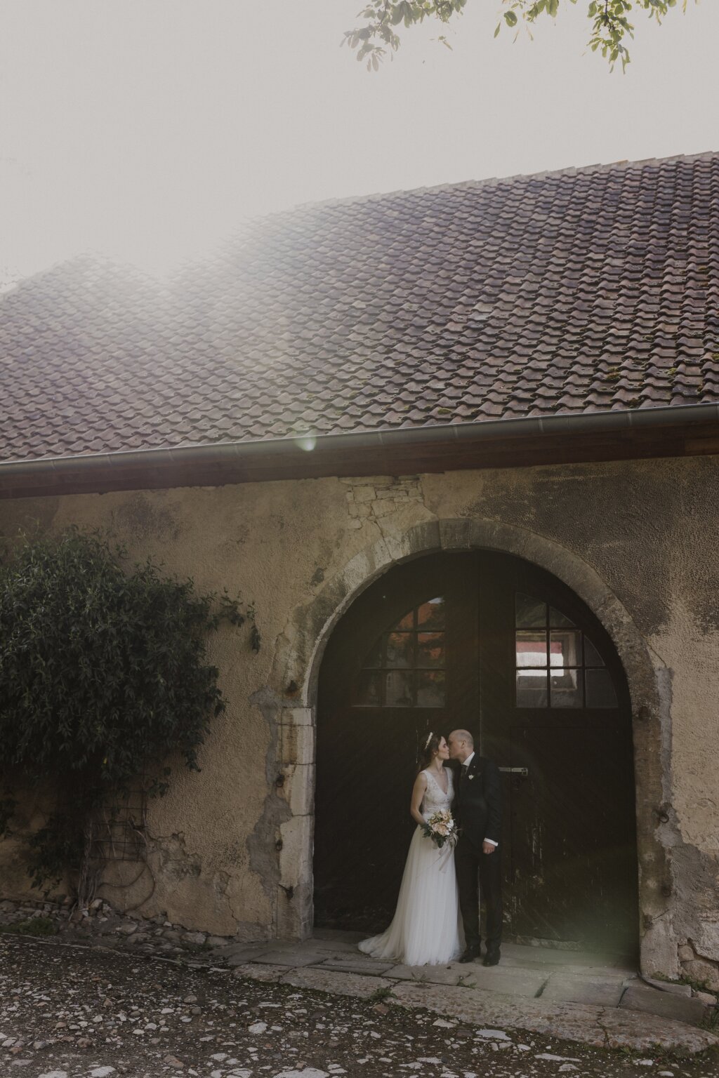 Ein Brautpaar küsst sich verliebt vor einer alten Scheunentür. Das Abendlich scheint über das Dach der Scheune und erzeugt tolle Lichteffekte.