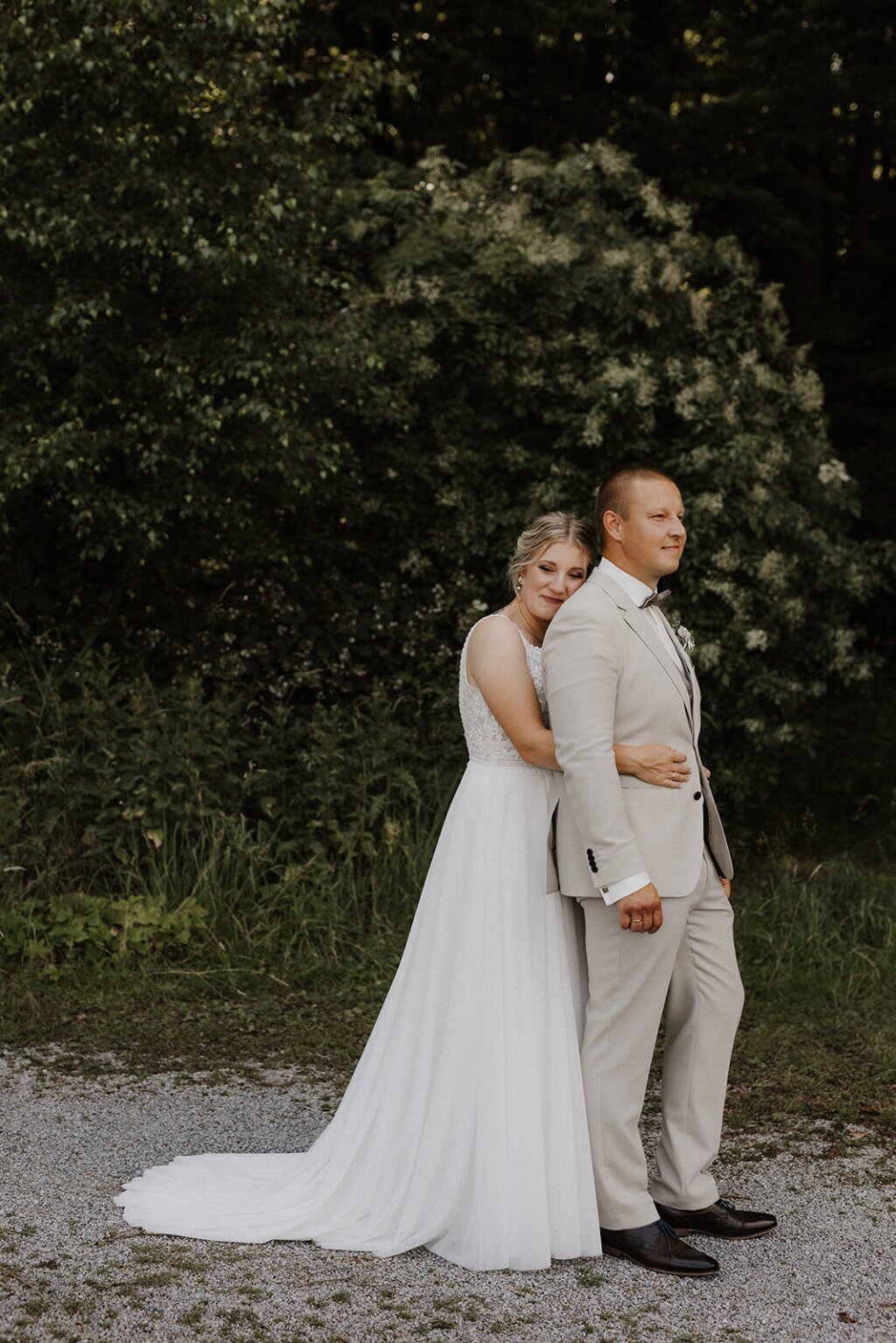 Ein Brautpaar steht am Waldrand auf einem Schottere. Im Hintergrund blüht der Holunder. Die Braut steht hinter dem Bräutigam und kuschelt sich an seinem Rücken.