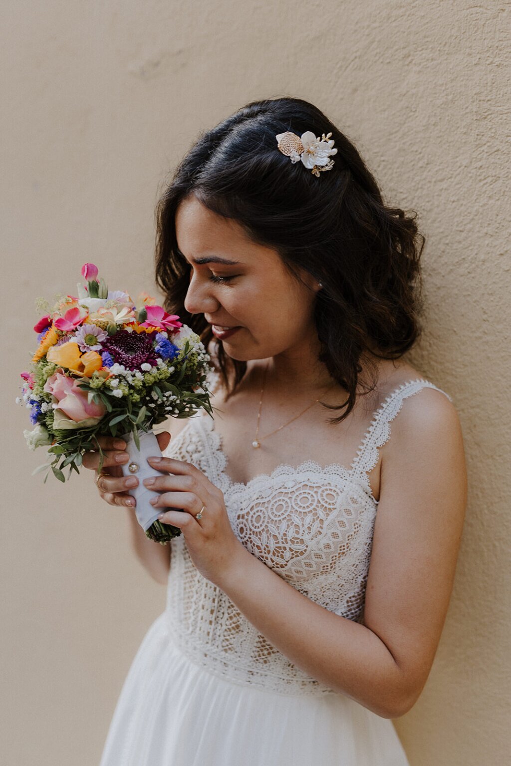Eine Braut in einem weißen Kleid lehnt sich gegen eine beige Wand und riecht an ihrem bunten Brautstrauß. Sie trägt eine schöne goldene Spange in ihren schwarzen Haaren.