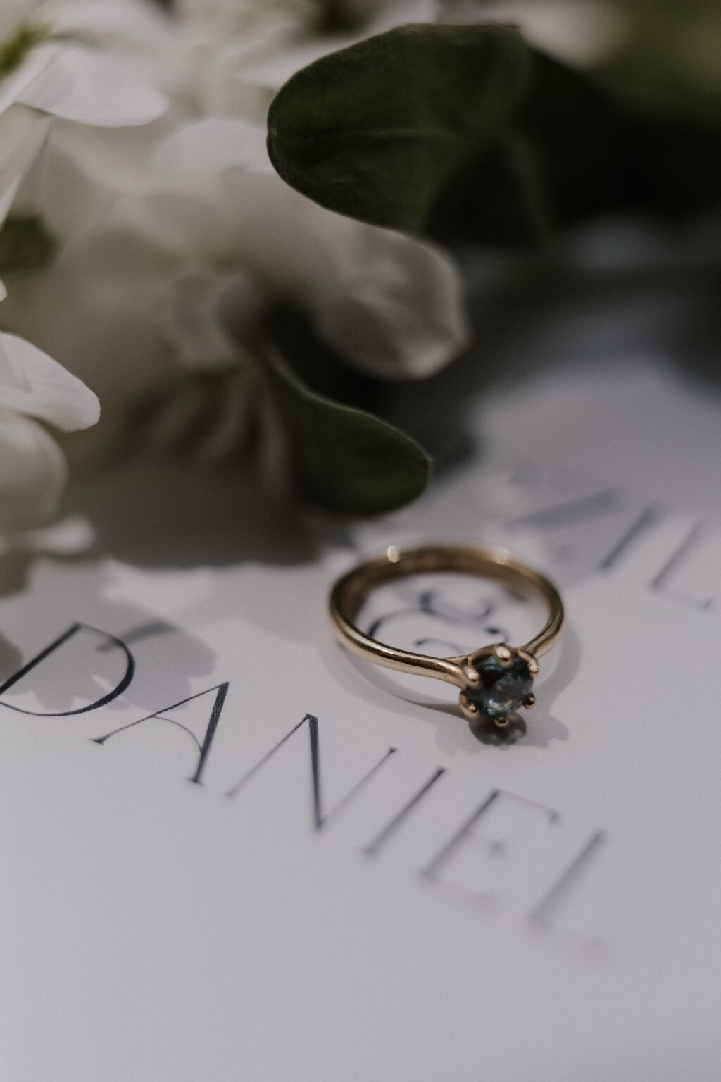 Ein goldener Verlobungsring mit eingefasstem blauen Stein liegt auf einem Stück Papier. Zu erkennen ist der Name Daniel. Neben dem Ring lassen sich Teile einer Blume erkennen.