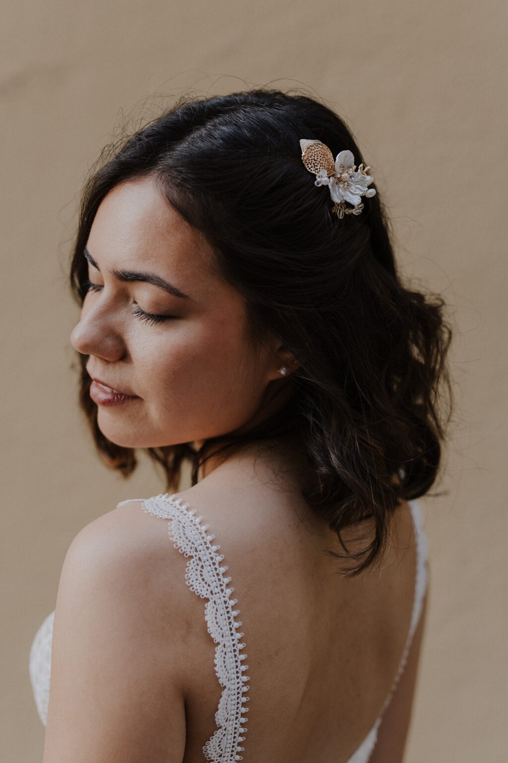 Eine hübsche Braut schaut mit geschlossenen Augen sanft über die Schulter. Ihre toll zurechtgemachten Haare und ihr elegantes Braut-Make-Up kommt gut zur Geltung. Sie trägt eine florale Haarspange mit goldenen Elementen.