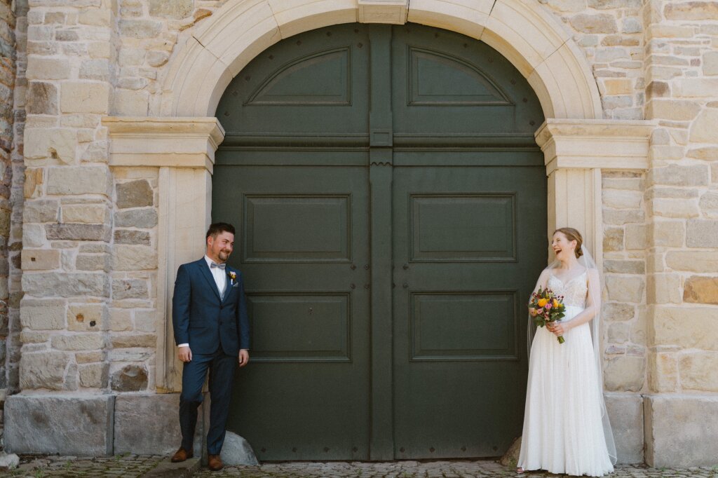 Ein lachendes Brautpaar steht an einer Wand gelehnt. Zwischen ihnen ist ein schönes altes Tor mit Rundbogen, sie lehnen an den Säulen der Tür und schauen sich an.