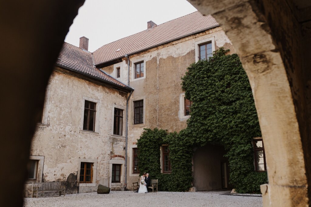 Ein Brautpaar sitzt im Innenhof eines alten Klosters auf einer Bank. Die Braut hat auf dem Schoß des Bräutigams Platz genommen und sie küssen sich. Die Fassade der Gebäude ist Teilweise offen und es sind alte Steine zu erkennen. Hinter dem Brautpaar ist die Wand mit Efeu bewachsen.