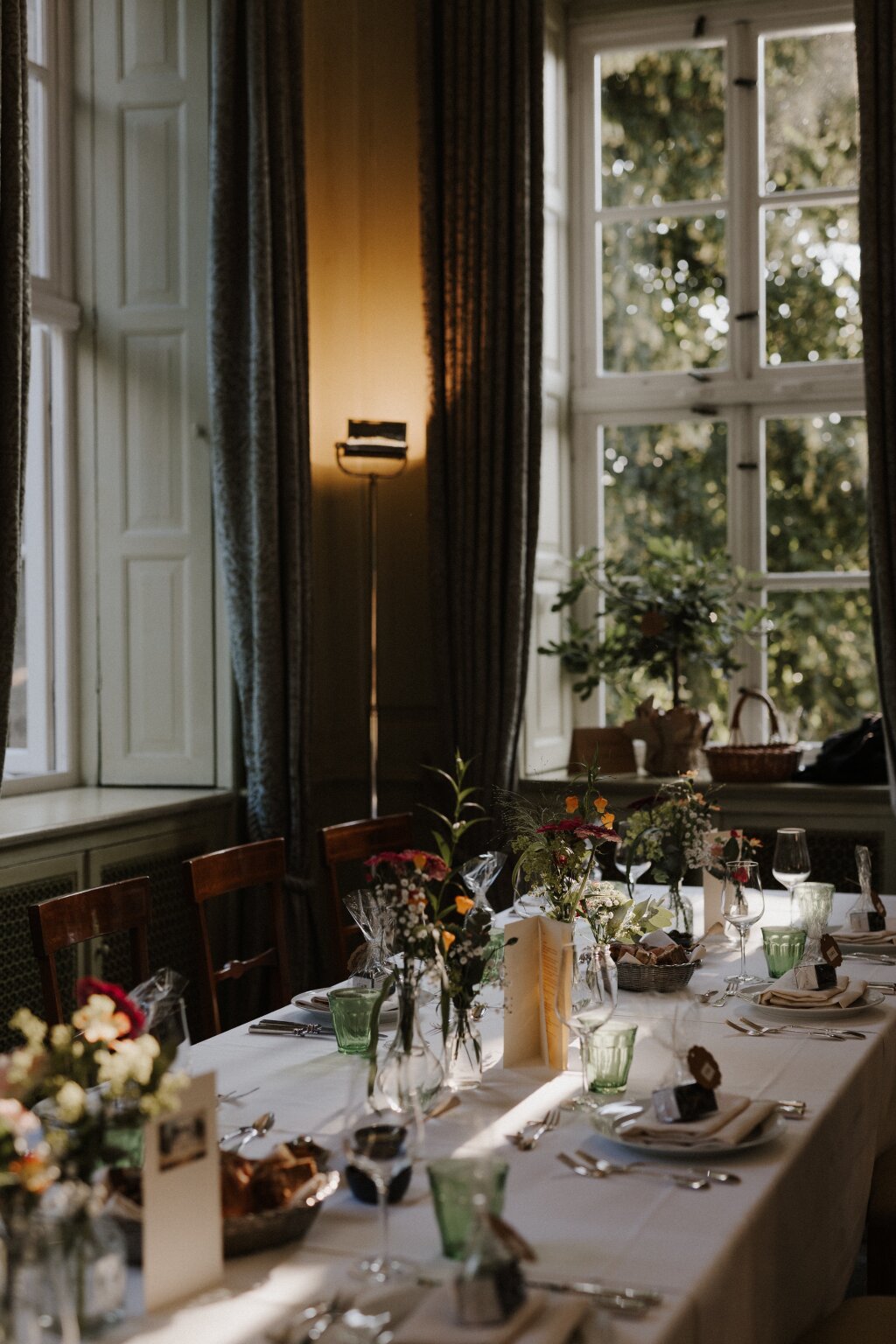 Ein festlich gedeckter Hochzeitstisch mit verschiedenen kleinen Vasen und Blumen. Eingedeckte Teller mit kleinen Gastgeschenken und Menükarten sind auf dem Tisch. Die Abendsonne fällt durch das Sprossenfenster hinein. In der Ecke steht eine Stehlampe und leuchtet die gemusterten, fließenden Gardinen an.
