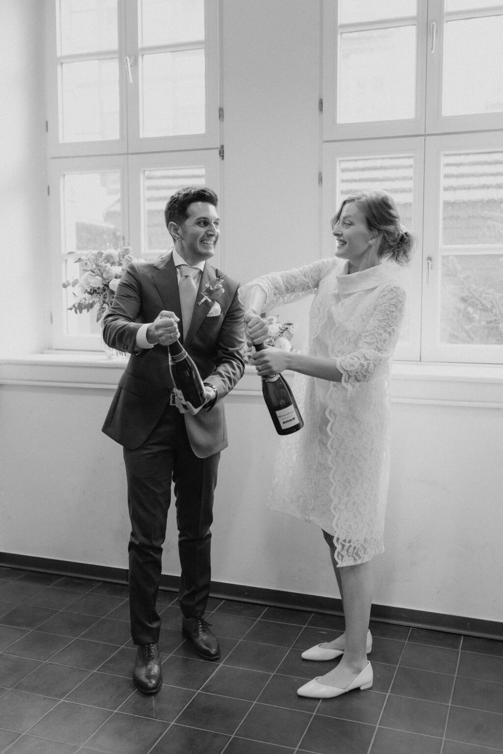 Ein Hochzeitspaar steht vor einer Fensterfront und öffnet jeweils eine Flasche Champagner. Dabei schauen sie sich lachend an.