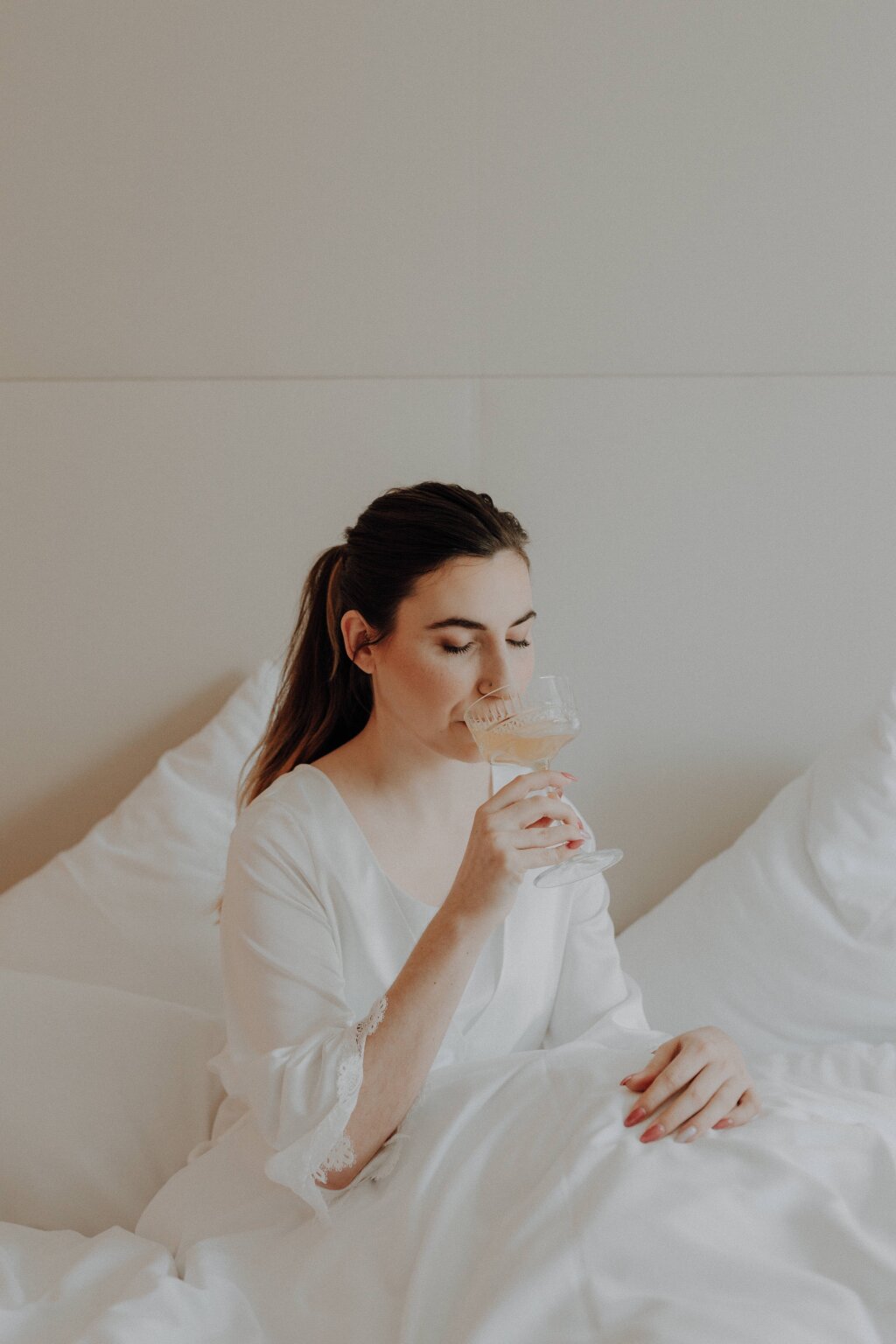 Eine Braut sitzt während des fertigmachens in einem weißen Hotelbett und trinkt elegant aus einem Champagnerkelch.